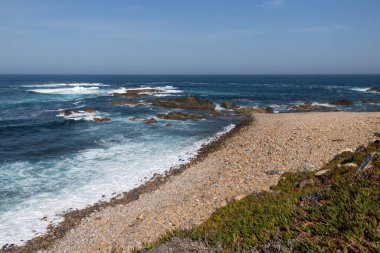 Arkaplanda kayalık dağlar ve su dalgalarıyla kaplı bir deniz kıyısı.