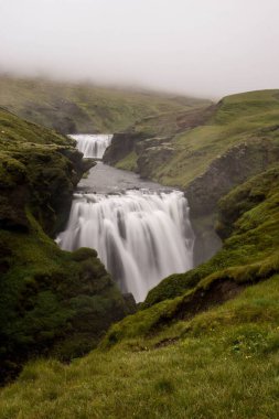 Gün boyunca bulutların altında bir şelalenin dikey görüntüsü.
