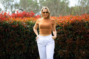 Kahverengi bluzlu ve beyaz pantolonlu genç bir İspanyol kadın parkta kameraya gülümsüyor.