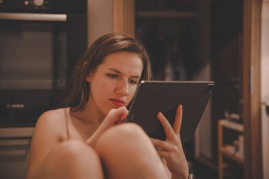 Genç beyaz bir kız mutfakta oturuyor ve tabletinde kitap okuyor.