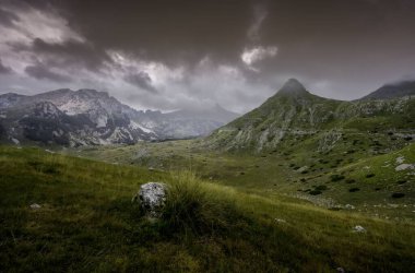 Bulutlu karanlık gökyüzünün altında yeşil yamaçlı dağların manzarası.