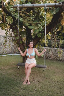 Tropik bir bahçede salıncakta sallanan genç bir kadının dikey çekimi..