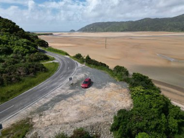 Dar yolda, kumlu bir alanda ve arkada tepeleri olan kırmızı arabanın manzarası.