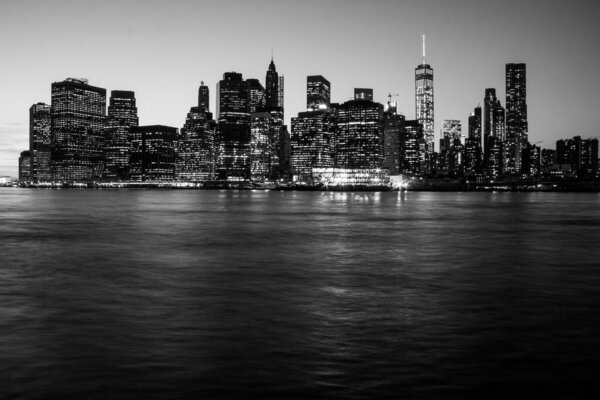 A grayscale of the glowing Manhattan cityscape with a river in the foreground at night