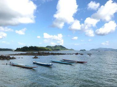 Huzurlu sularda, parlak bulutlu gökyüzünün altında yeşil tepelerle çevrili bir sıra uzun kayık.