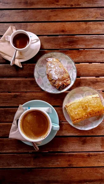 扁平的杏仁羊角面包和膨胀起来的金枪鱼糕点放在一张木制桌子上 配上一杯浓缩咖啡和黑咖啡 作为简单的法国早餐 — 图库照片