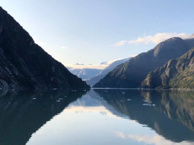 Güneşli bir günde sulara yansıyan büyük dağların olduğu sakin bir göl.