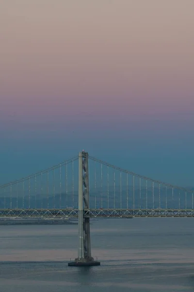 位于加利福尼亚州旧金山的奥克兰湾大桥在粉色和蓝色天空下的水面上垂直拍摄的照片 — 图库照片