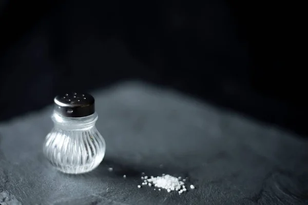 玻璃盐瓶的特写镜头和散布在灰蒙蒙表面的一些盐粒 — 图库照片