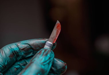 Kana bulanmış neşter tutan yeşil plastik eldivenli bir el.