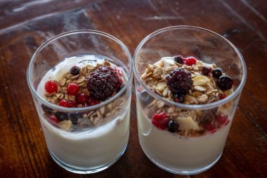 Tahta bir masada iki bardak yoğurt, granola ve böğürtlen.