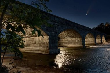 Bir taş köprünün alçak açılı görüntüsü ve gökyüzünde gece boyunca görünen bir kuyruklu yıldız.