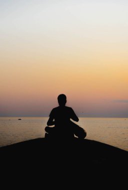 Gün batımında deniz kıyısında meditasyon yapan bir insanın siluetinin dikey görüntüsü.