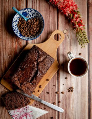 Kahveli kekin üst açılı bir resmi, ahşap mutfak tahtasında servis ediliyor, bir kase kahve çekirdeği ve bir fincan kahve ile birlikte.