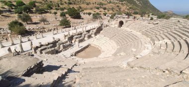 Türkiye 'nin Efes kentinin tarihi bir simgesi olan küçük bir tiyatro, güneşli bir günde arka planda alçak bitki örtüsü bulunuyor.