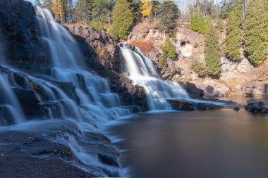 Güneşli bir günde yeşil ağaçlarla çevrili çağlayan bir çeşmenin uzun pozu.