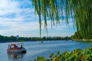 Ağaçlarla ve bitkilerle çevrili güzel bir doğa sahnesi. Gündüz pedallı teknelerde insanlar var.