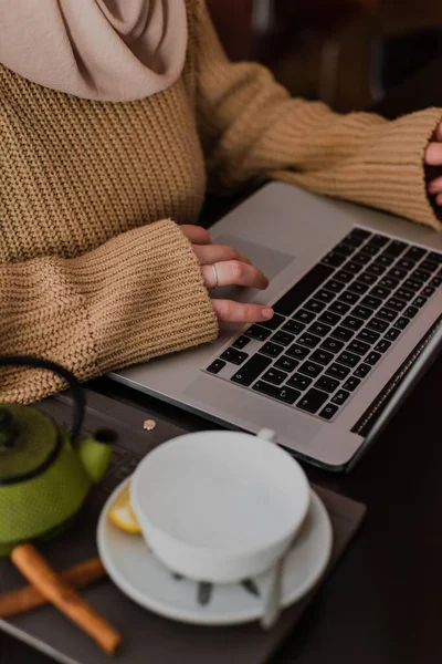 カフェで茶色いセーターを着てノートパソコンの上でティーカップと鍋で働いていた女の子が — ストック写真