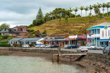 Mangonui 'de yeşil bir tepenin yakınındaki dükkanların önündeki rıhtım yolunda park edilmiş arabaların manzarası.