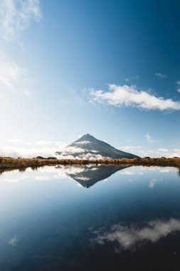 Etrafında tepeler ve bulutların yansıması olan bir gölün manzaralı dikey görüntüsü.