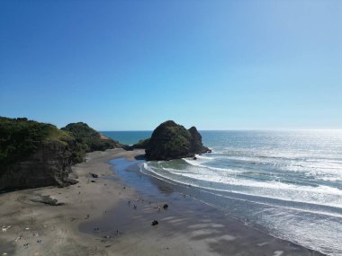 Sakin bir deniz ve mavi gökyüzü günüyle kumsaldaki insanların insansız hava aracı görüntüsü. Arka planda kayalık bir uçurum.