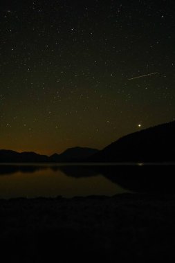 Geceleri büyüleyici yıldızlı gökyüzünün altında tepelerle çevrili sakin bir gölün manzaralı dikey görüntüsü.