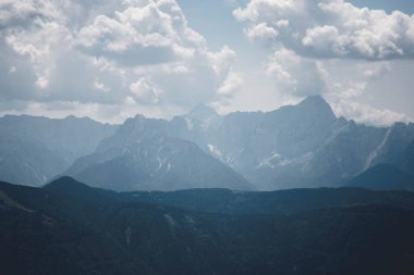 Büyük kayalık dağların nefes kesici manzarası çimenli tarlaların üzerinde beliriyor ve onları parlak bulutlu bir gökyüzünün altında gölgeyle kaplıyor.