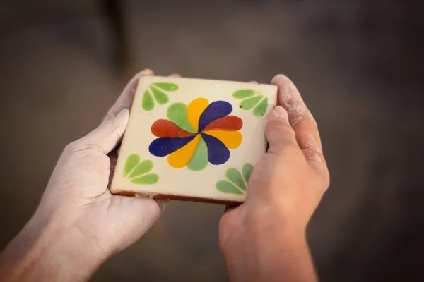 A closeup of dusty human hands holding beautiful square Mexican ceramic tile with floral patterns