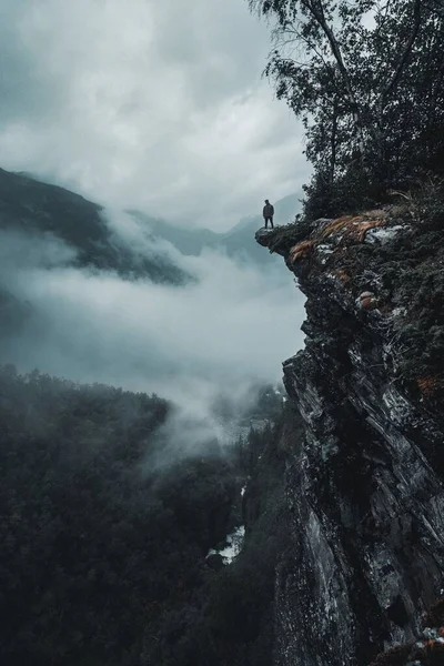 一个男子在面对雾蒙蒙的森林的岩石悬崖边缘上的垂直无人机射击 — 图库照片