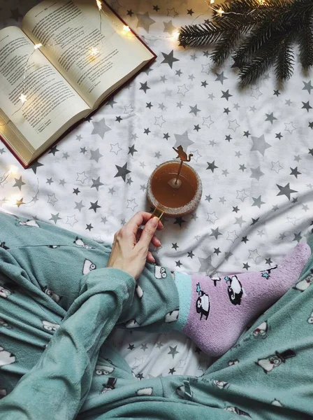 本とクリスマスライト付きの柔らかいベッドの上でお茶のカップを持つ女性の手の垂直上のビュー — ストック写真