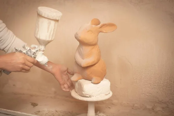 スタジオのスタンドでかわいい陶器のウサギに白い塗料を吹きかける人間の手 — ストック写真