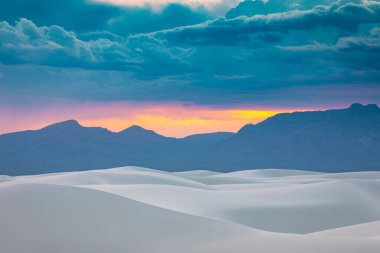Gün batımında bulutlu bir gökyüzünün altında dağların ufuk çizgisi ve gri kum tepeleri
