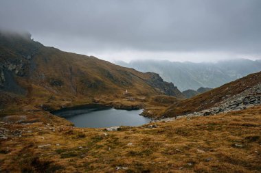 Romanya 'daki güzel küçük Capra Gölü kasvetli gökyüzünün altındaki büyük dağlarla çevrili.