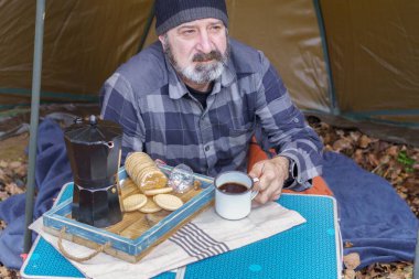 Sakallı ve şapkalı bir adam kış çadırında kahve içiyor.