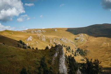 Mavi bulutlu gökyüzünün altındaki kayalık tepelerin ve tarlaların kırsal bir manzarası.