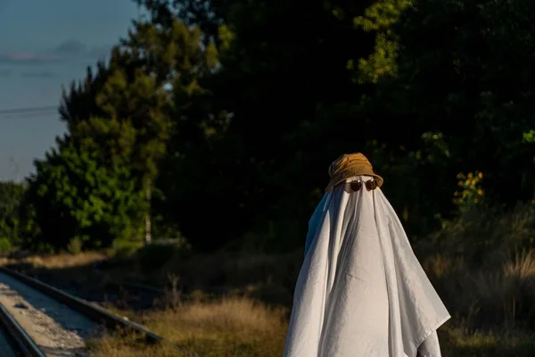 Osoba Nakryta Białym Prześcieradłem Okularach Przeciwsłonecznych Kapeluszu Wyglądem Ducha Ogrodzie — Zdjęcie stockowe