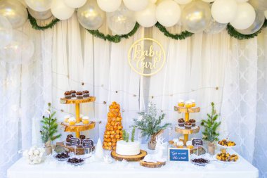 Bebek partisinde duvarında yeşil süslemeler ve beyaz balonlar asılı duran bir masada pasta ve şekerler.