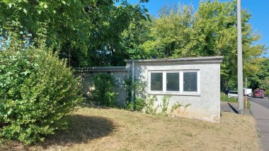 Güneşli bir günde ağaçlar ve çalılarla çevrili tek katlı bir bina.