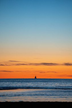 Gün batımında turuncu gökyüzüne karşı tek bir yelkenli teknesinin dikey çekimi.