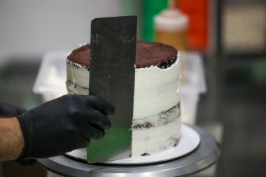 Pasta şefi tereyağlı, uzun cilindrik kremalı pastayı pürüzsüz yapmak için spatula kazıyıcısı kullanıyor. Düğün pastası hazırlığı.