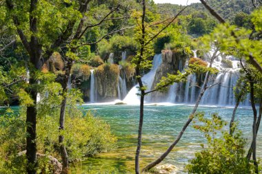 Manzaralı şelale, yeşilliklerle çevrili berrak mavi suya dökülüyor..