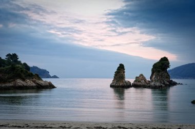 Bulutlu gökyüzünün altında sakin okyanus kıyısında kayaların manzarası.