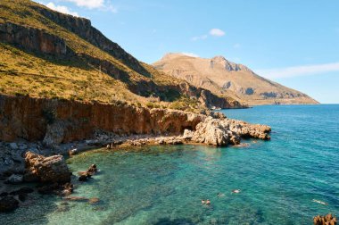 Berrak turkuaz deniz, engebeli kayalık kıyı şeridi parlak mavi gökyüzü altında, yükselen uzak dağlar..