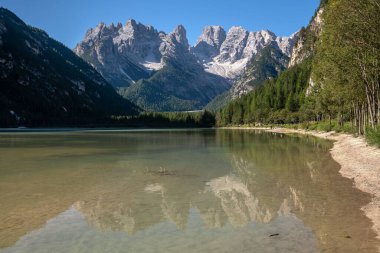 Mavi gökyüzünün altındaki görkemli dağları yansıtan berrak göl manzarası.