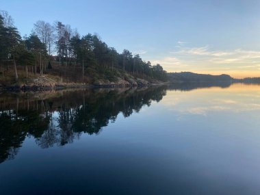 Uzun yeşil ağaçlar ve kayalık bir kıyı şeridi sakin bir gölün cam ve sakin yüzeyinde mükemmel bir şekilde yansıtılır..
