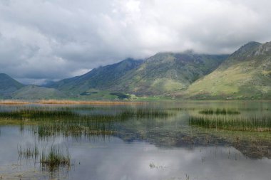 Bereketli yeşil tepelerin yer aldığı manzara bulutlu gökyüzünün altında sakin sulara yansıyor