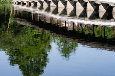 Yeşil ağaçlarla çevrili sakin suya yansıyan köprü.