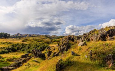 Yüksek irtifada yuvarlanan tepeler, kayalık çıkıntılar ve dramatik, bulutlu bir gökyüzü altında antik taş kalıntıları..