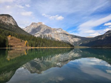 Mavi bulutlu gökyüzünün altında berrak göl sularına yansıyan dağların manzarası