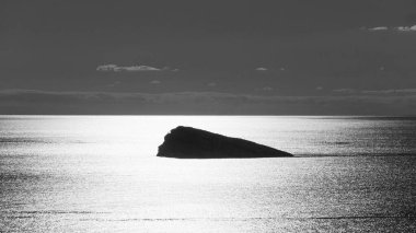 Sakin okyanus suyundan yükselen kayalık ada siluetinin siyah beyaz fotoğrafı